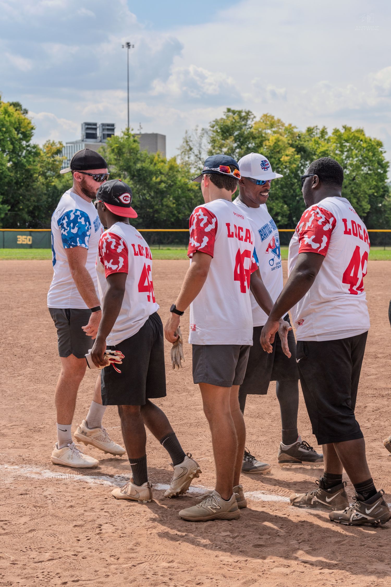 Group of softball players in matching white LUCA jerseys standing on a baseball diamond during a sunny outdoor game.