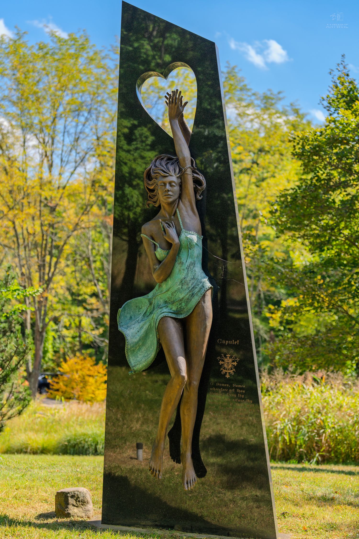 Bronze sculpture of a dancing woman with a heart motif on a dark obelisk in a lush green garden.