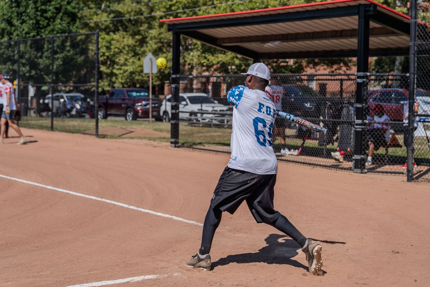A softball player in a white jersey swings a bat on a dirt field during an outdoor recreational game.