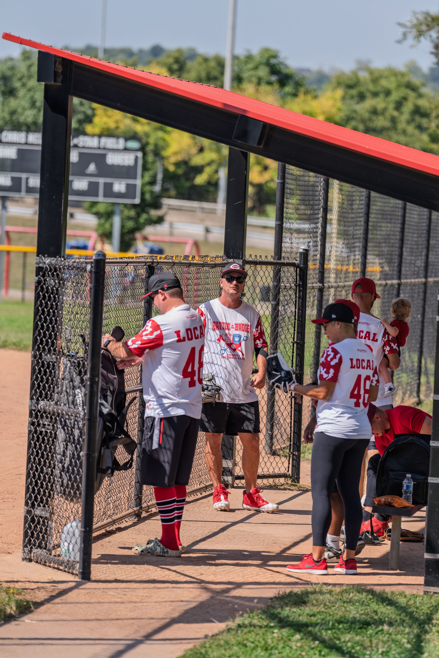 Baseball team players in red and white uniforms relaxing in dugout during sunny outdoor game day.