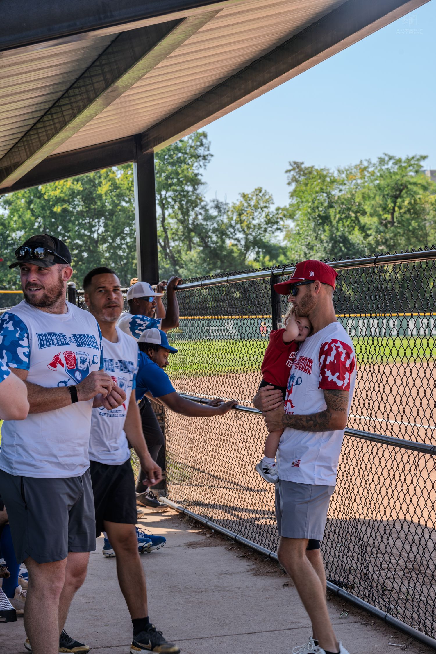 Players and coaches gather in a dugout during a recreational softball game on a sunny day.