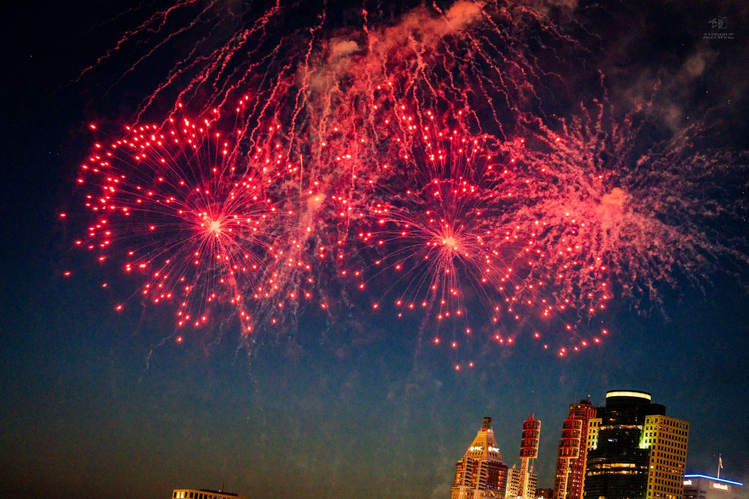 Vibrant red fireworks exploding over a city skyline against a dark blue night sky.