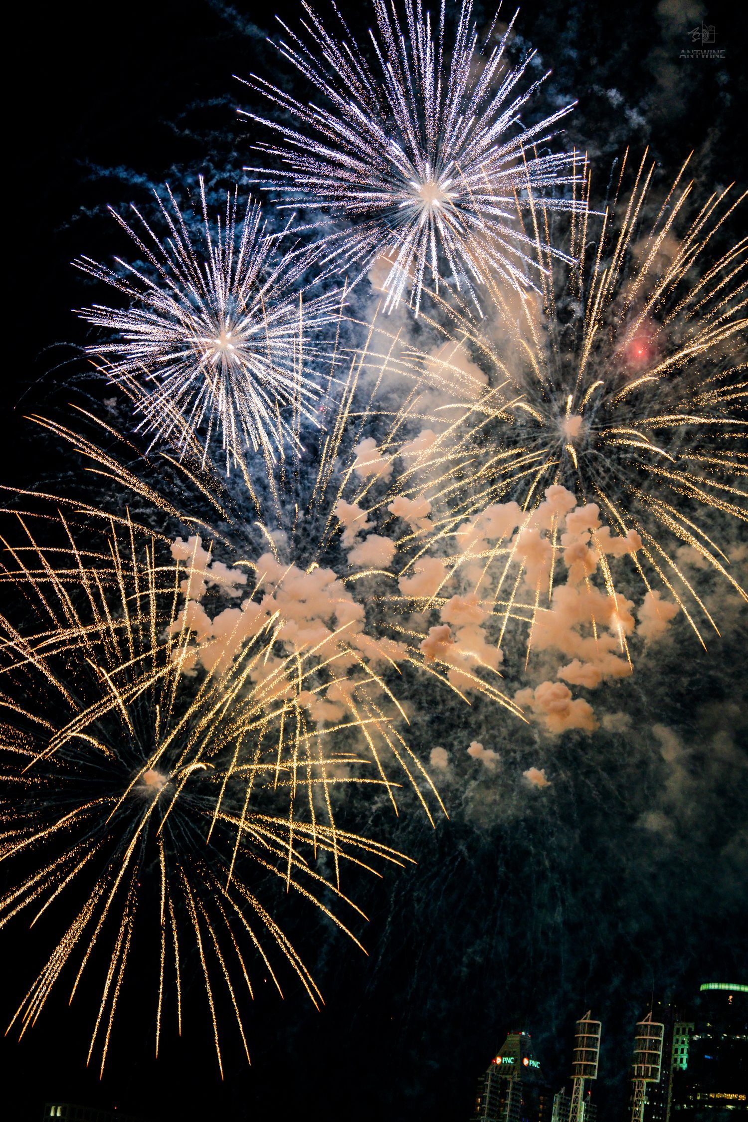 Spectacular multi-colored fireworks display with gold, white and blue bursts illuminating a smoky night sky over a city.