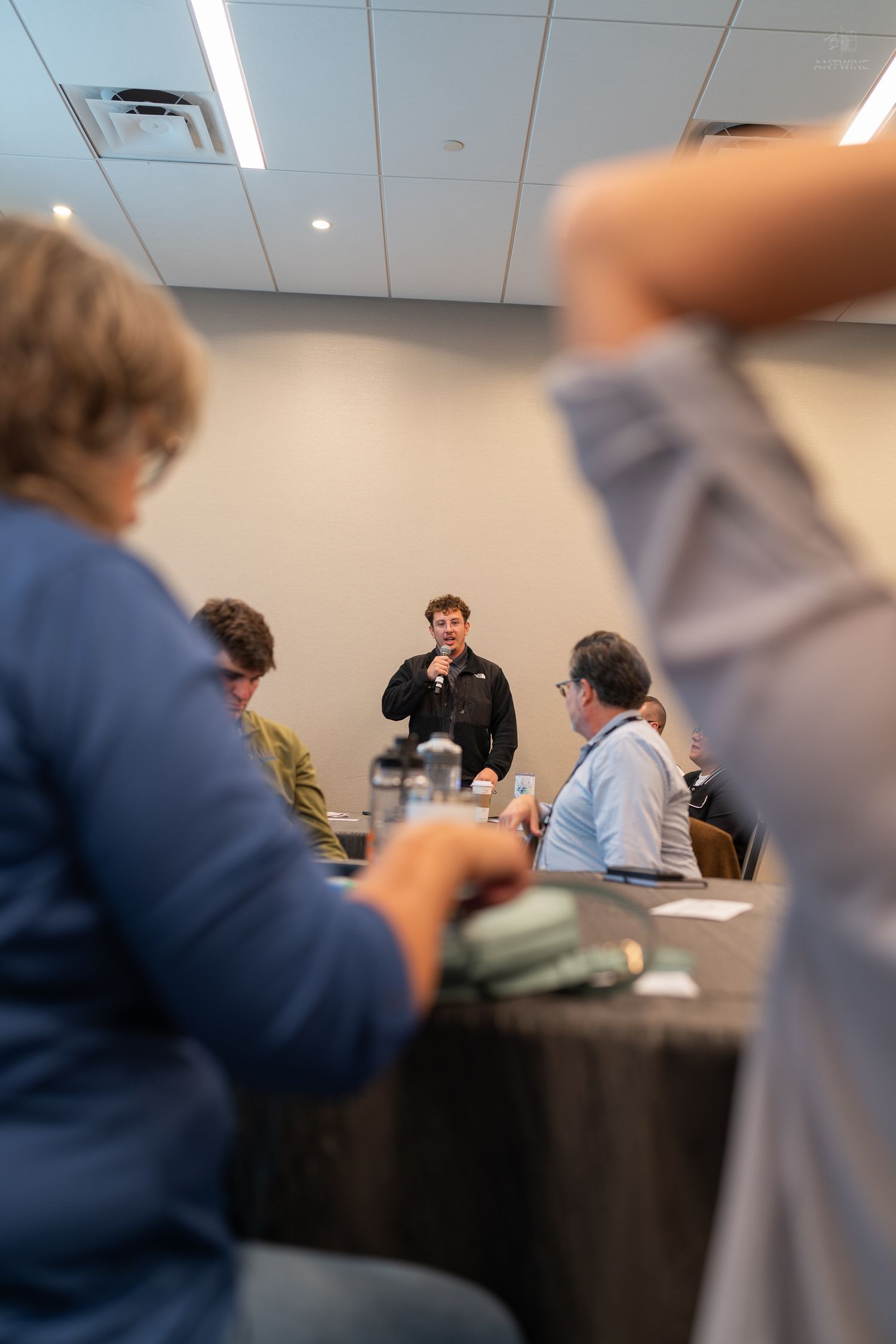 A speaker addresses a small group at a conference room meeting while an audience member raises their hand in the foreground.