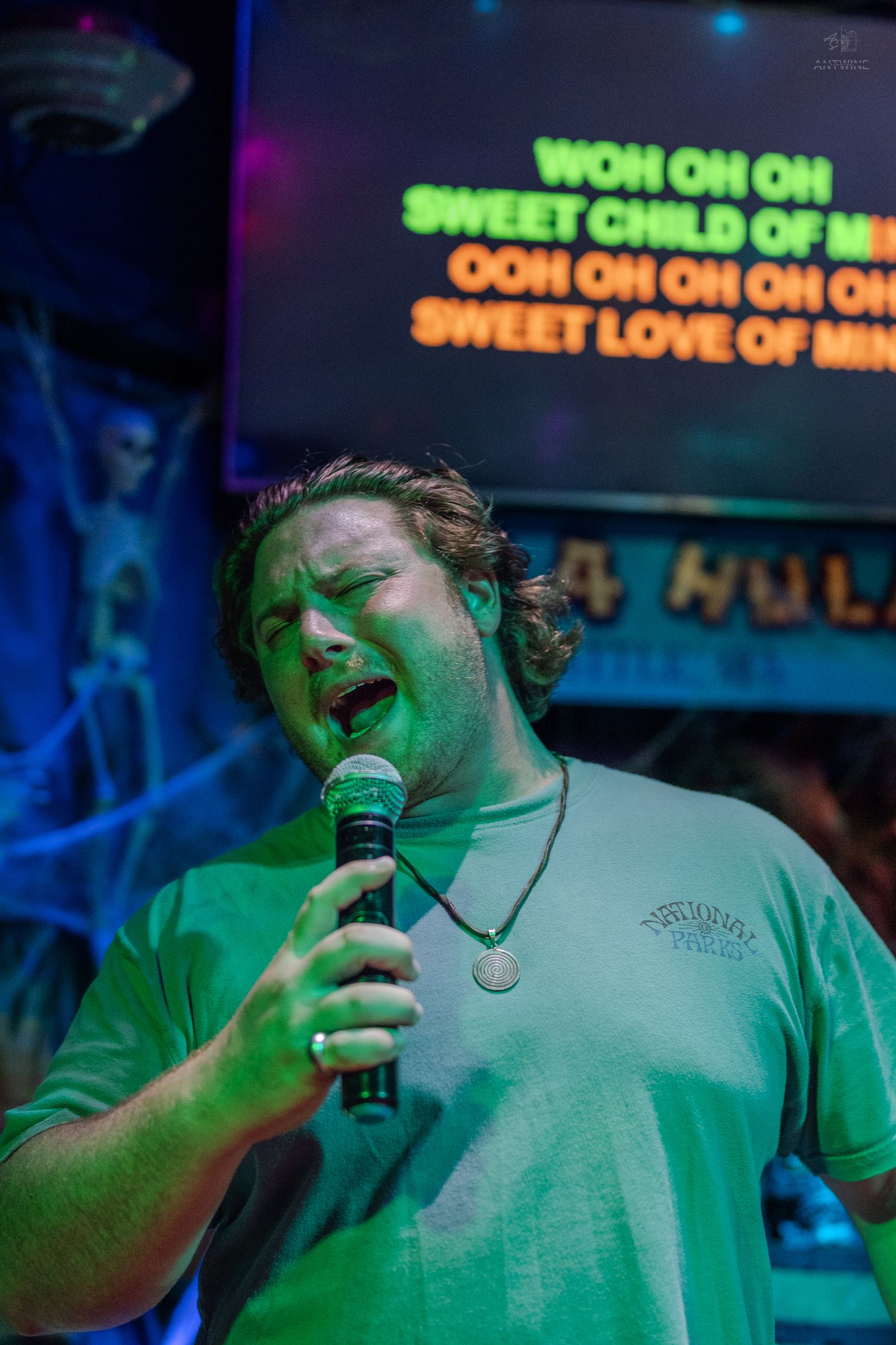 Man singing karaoke to Sweet Child of Mine at Hula Hula bar in Seattle under green stage lighting.