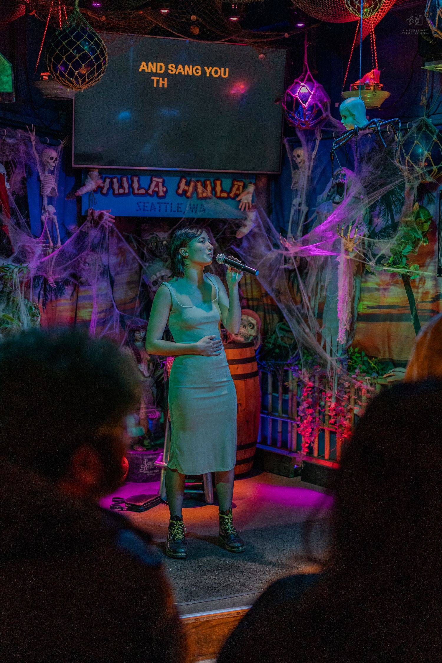 Woman singing karaoke on stage at a Halloween-decorated bar with spooky cobwebs and skeleton decorations.
