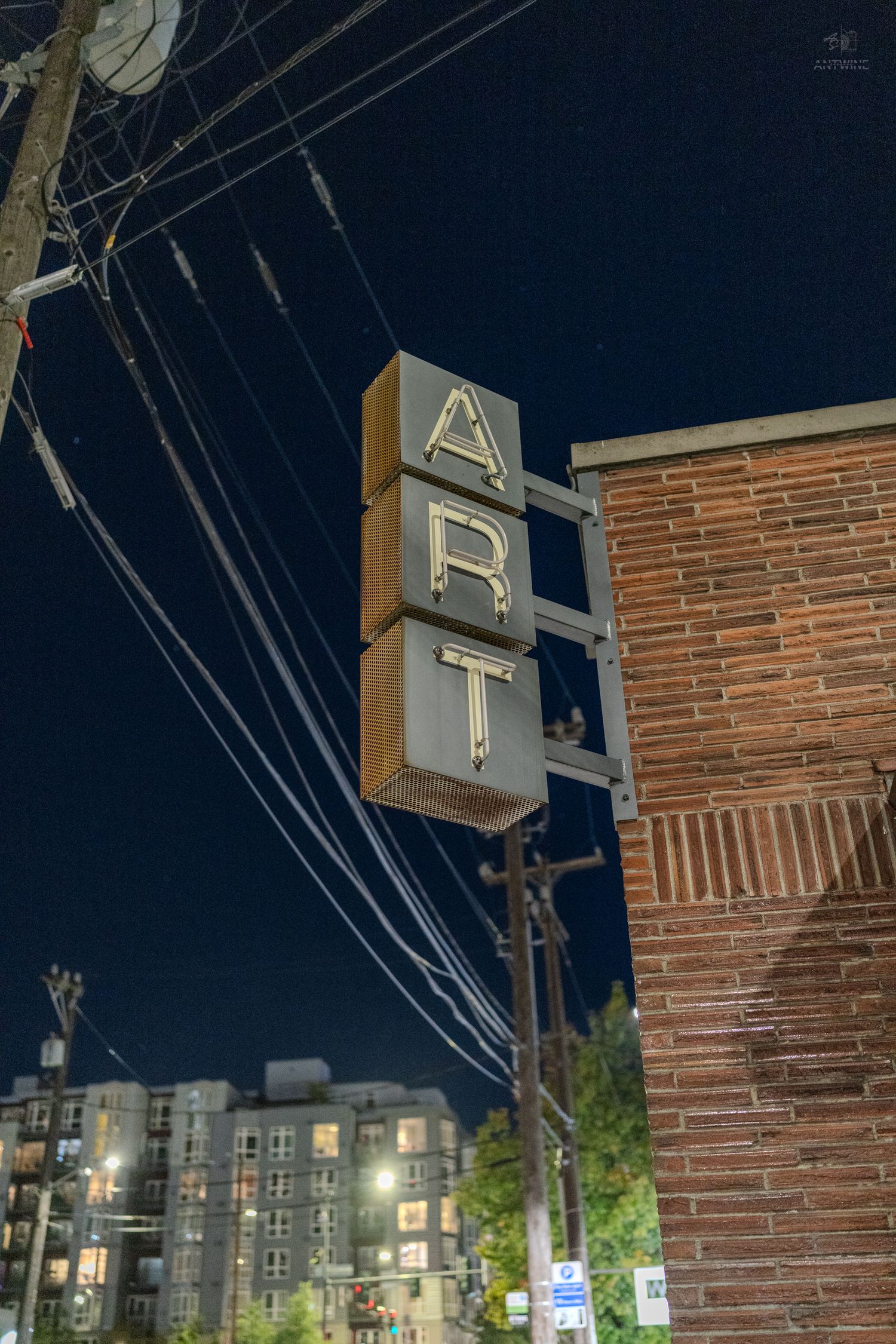 Vintage illuminated ART sign mounted on brick building corner at night with power lines and city skyline backdrop.