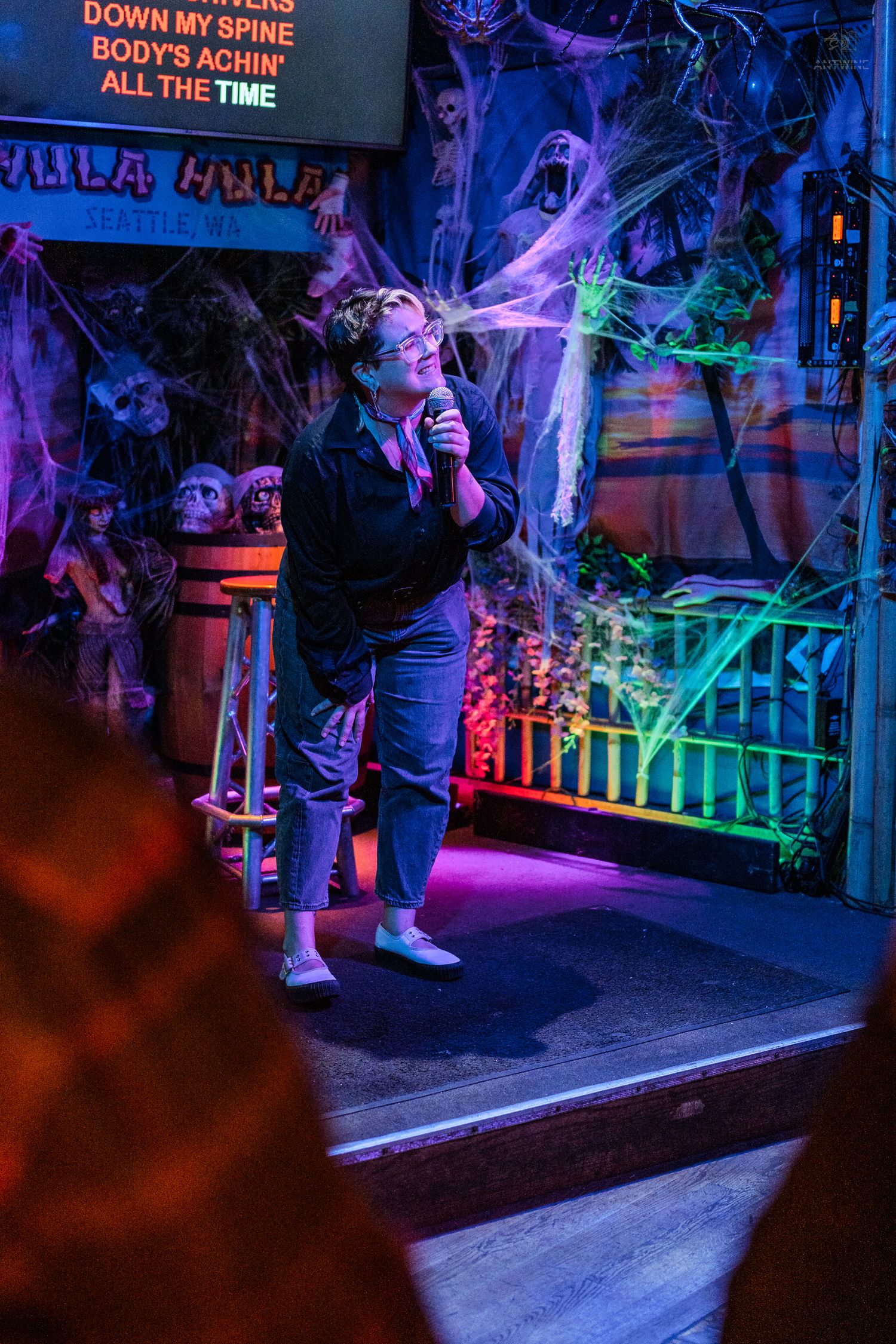 Person performing karaoke under colorful neon lights on spooky Halloween-themed stage at Hula Hula Seattle.