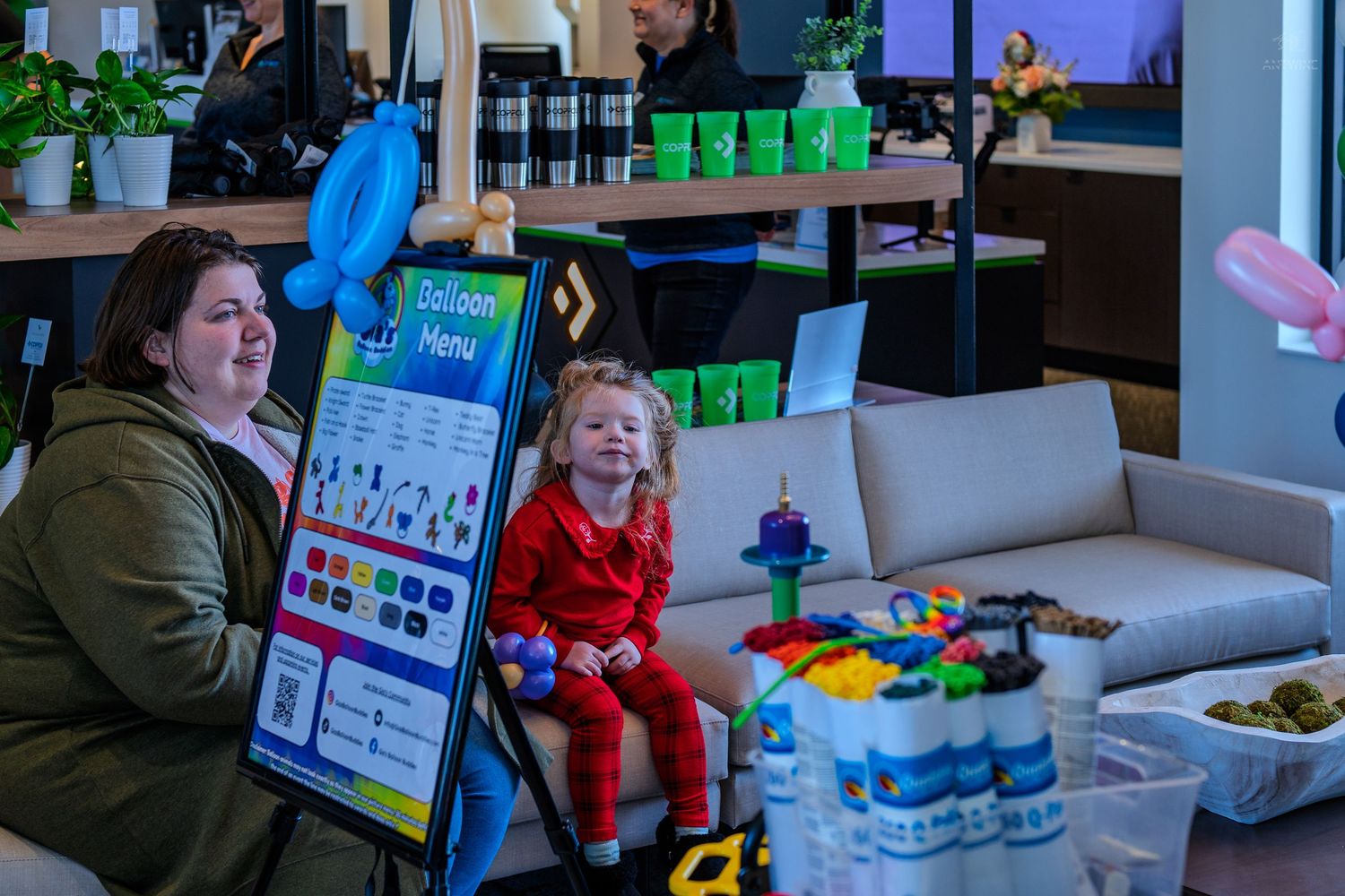 Mother and toddler sit together near a balloon menu display board at a festive indoor children's event.