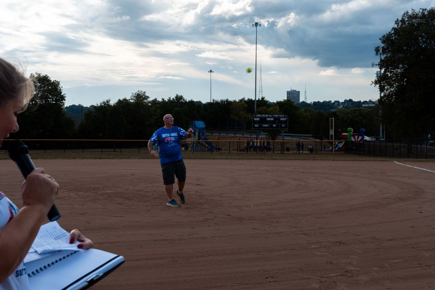 A man in a blue shirt throws a ball on a sandy softball field under cloudy skies at dusk.