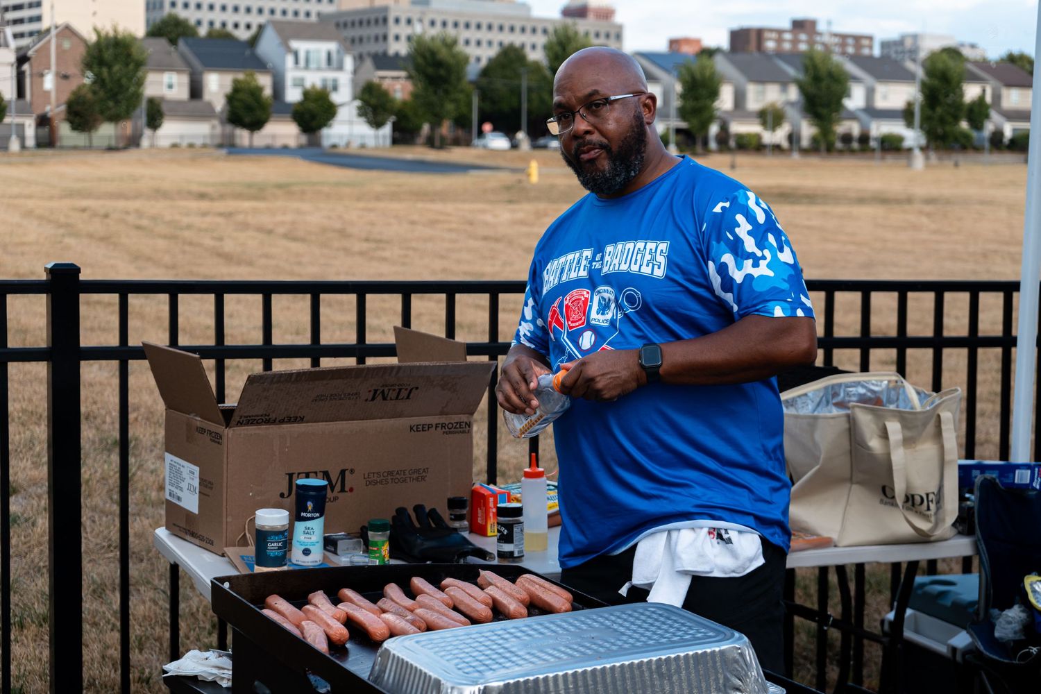 A man in a blue shirt grills hot dogs at an outdoor Battle of Badges charity softball event.