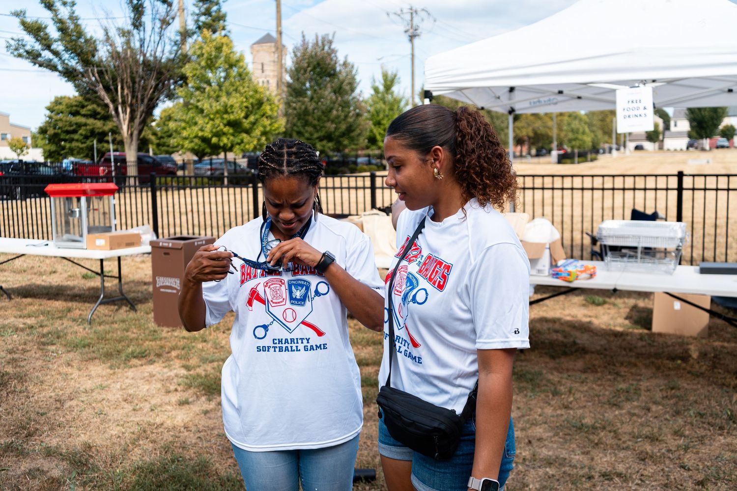Two women in white charity softball game shirts examine a medal outdoors at a community event.