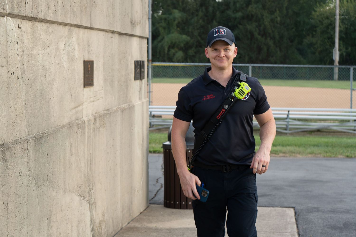A man in black uniform and cap stands confidently near a concrete wall by a baseball field.