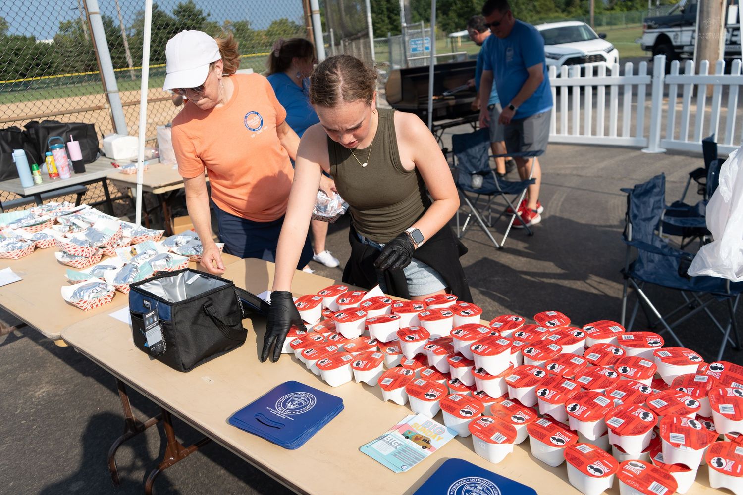 Two volunteers in gloves prepare red and white cups of food at an outdoor event table on a sunny day.