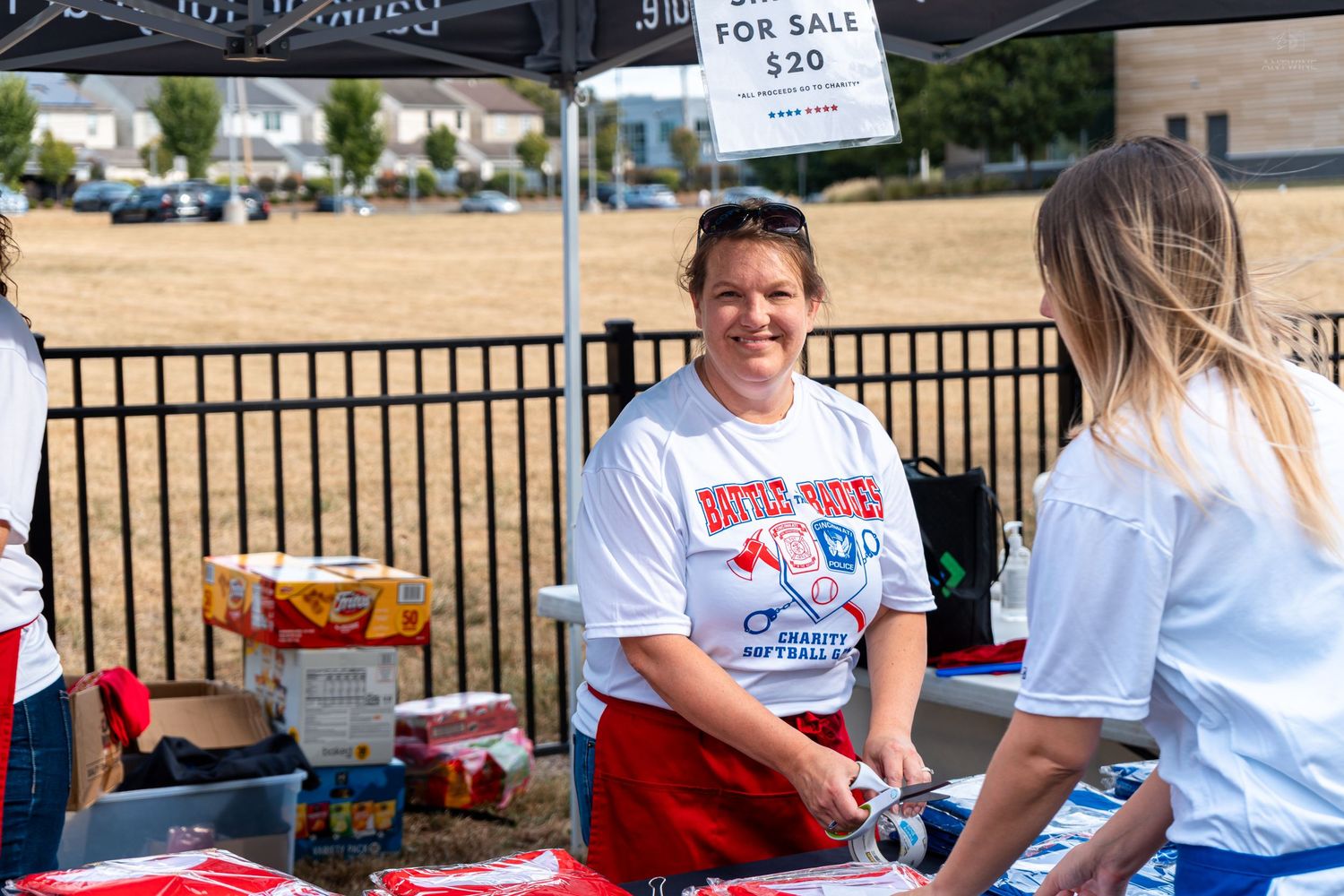 Two women smile at an outdoor market booth with a FOR SALE $20 sign, wearing white event t-shirts on a sunny day.