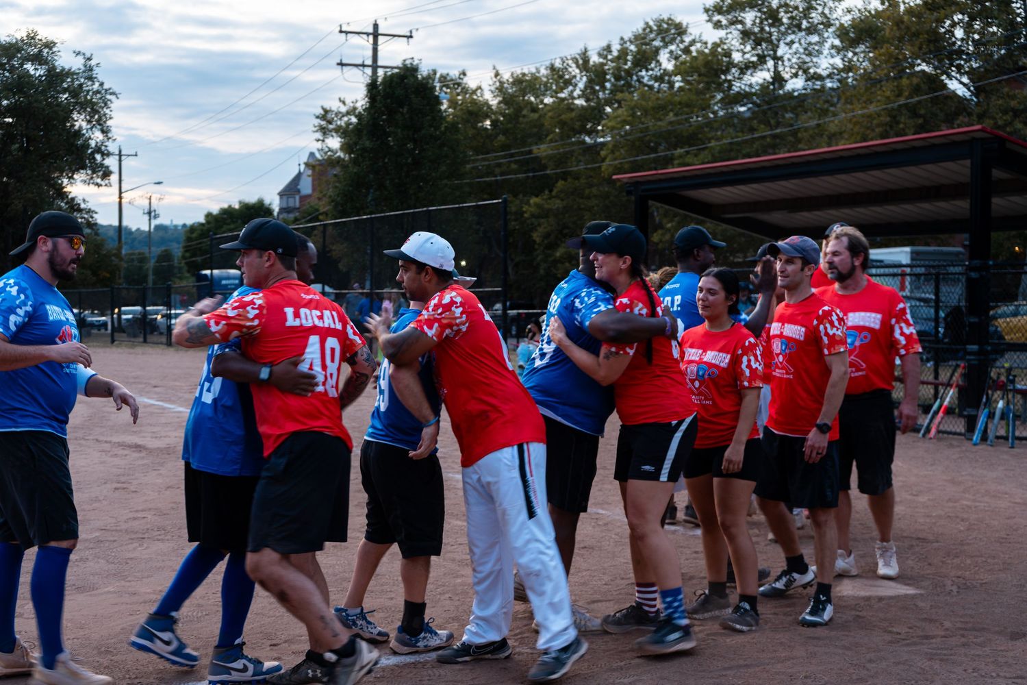 Softball teams in red and blue uniforms shake hands after a game on an outdoor field at dusk.