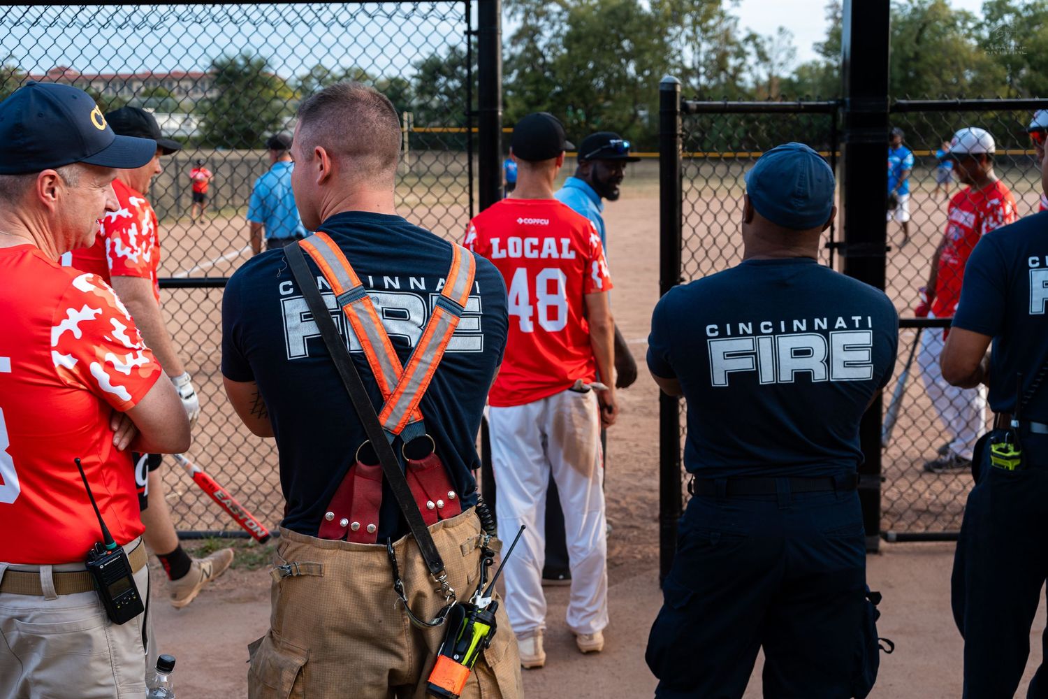 Cincinnati Fire department members in red jerseys and black uniforms gather at a baseball field fence during a local community event.