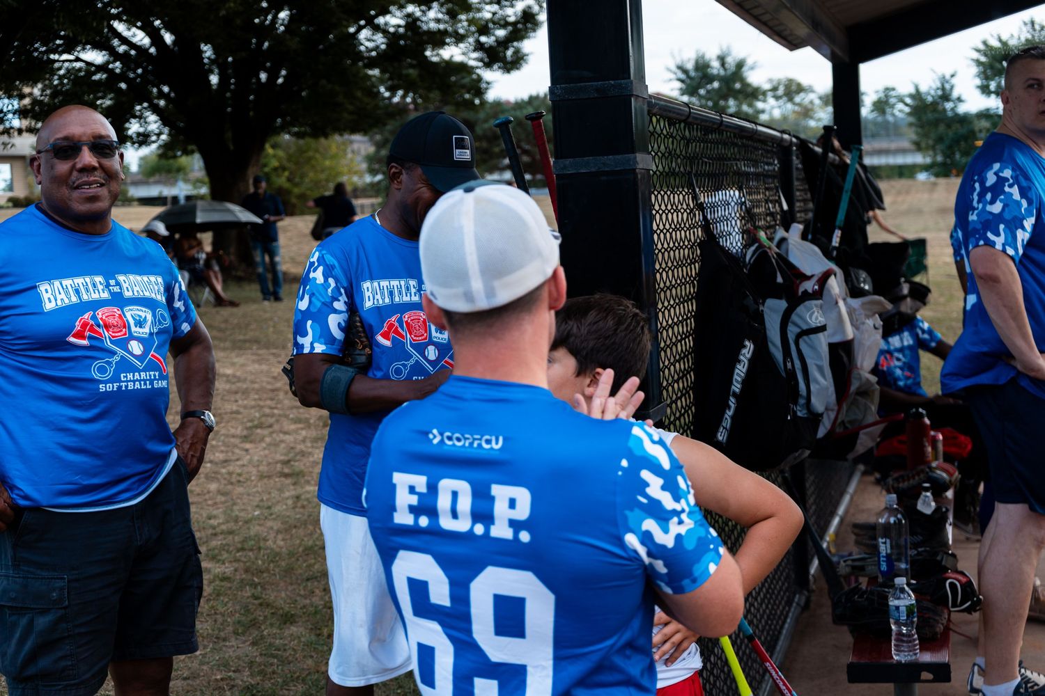 Players wearing blue F.O.P. 69 jerseys stand near a chain-link fence dugout at a softball game.