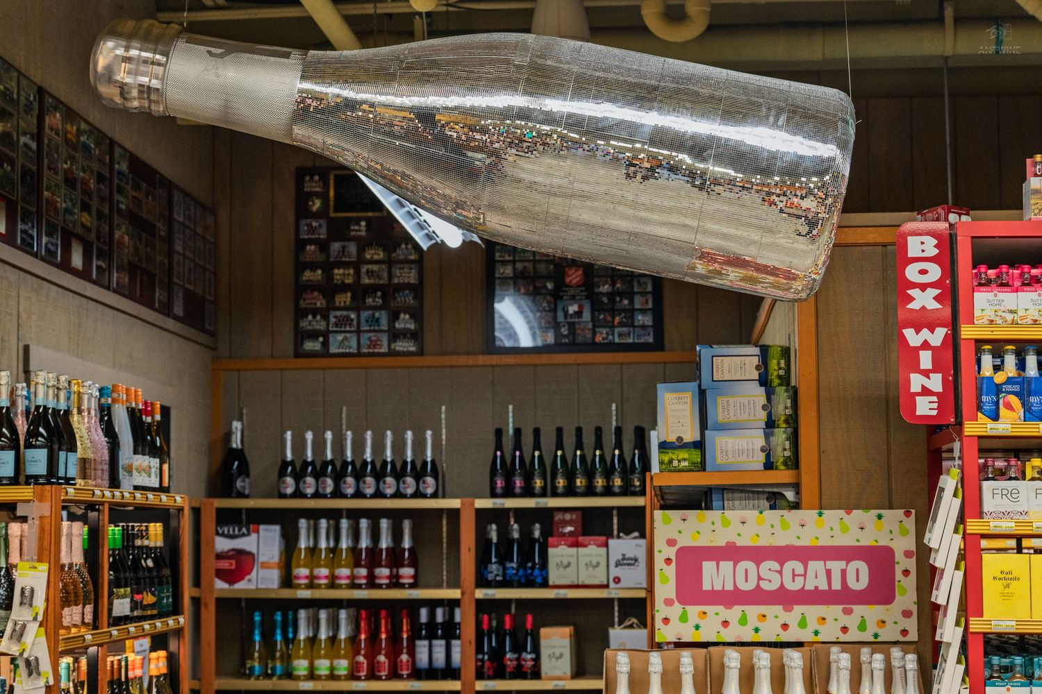 Large decorative silver bottle suspended above wine shelves with Moscato sign in a liquor store.