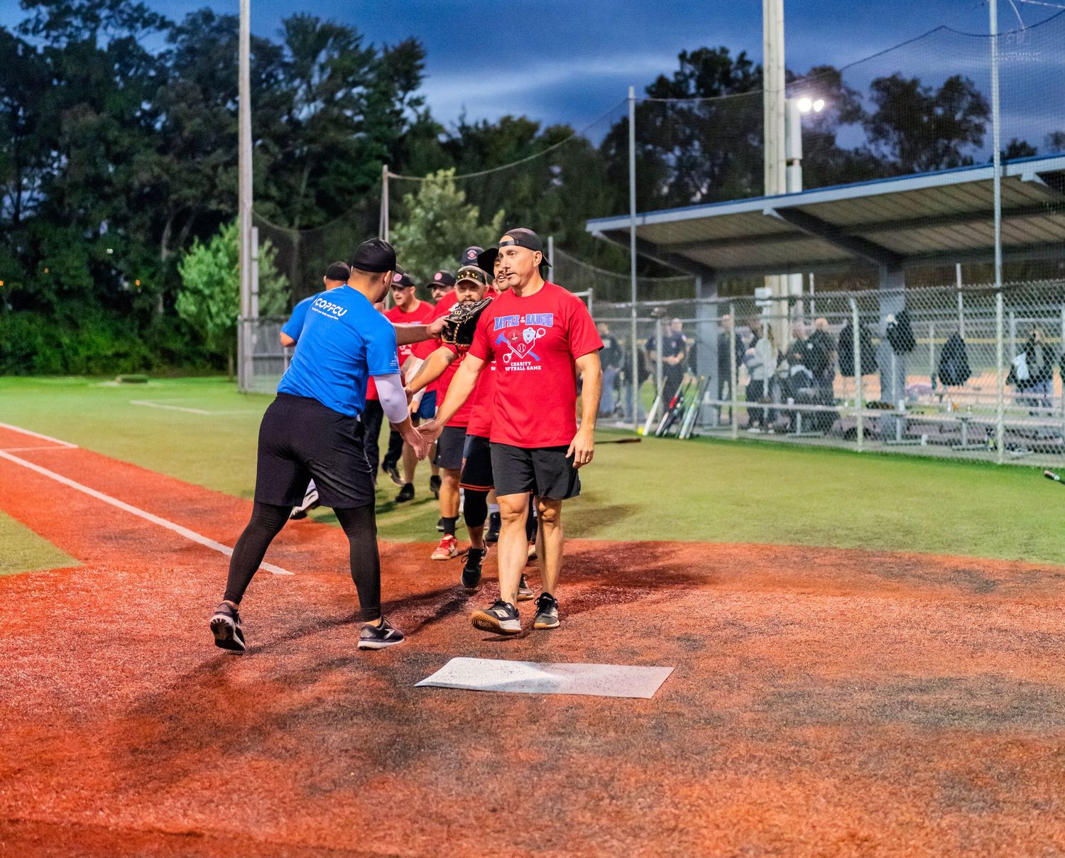 Players shake hands at home plate on a softball field at dusk, with bleachers and trees visible in the background.