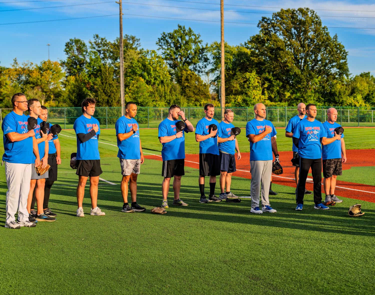 Group of players in blue shirts stand at attention on a turf field during a softball game ceremony.