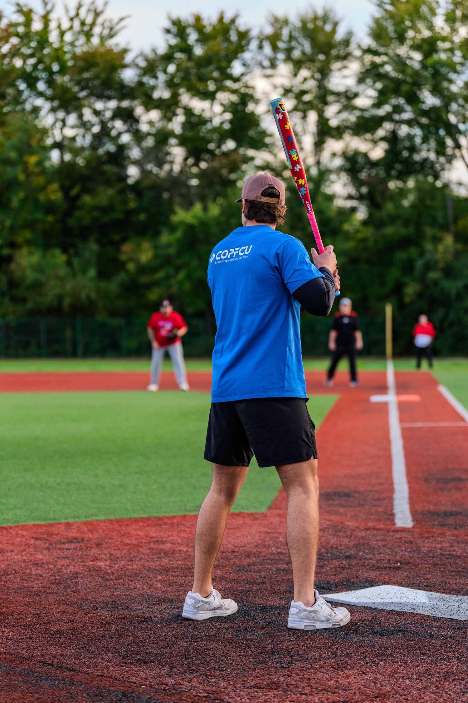 Male softball player in blue COOFCU shirt stands at home plate ready to bat on red turf field.