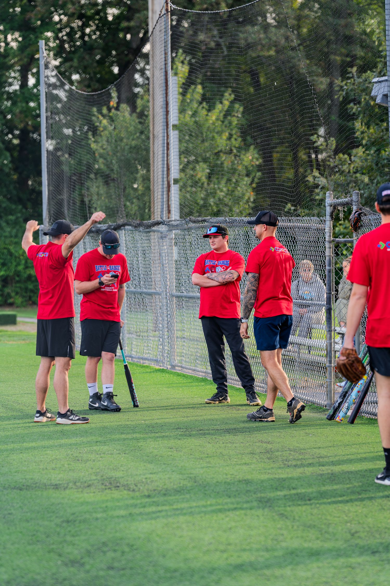 A group of softball players in red shirts gather near a chain-link fence during a warm evening game.