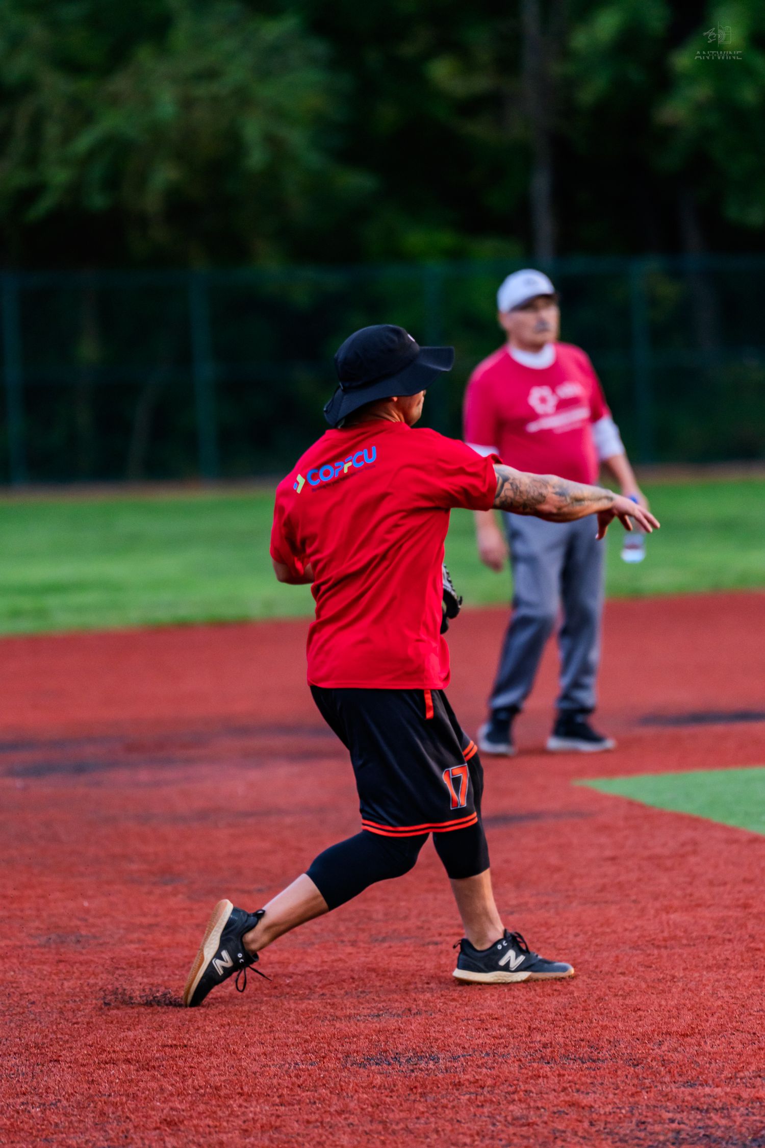A softball player in a red jersey and black pants throws the ball on a red turf infield.
