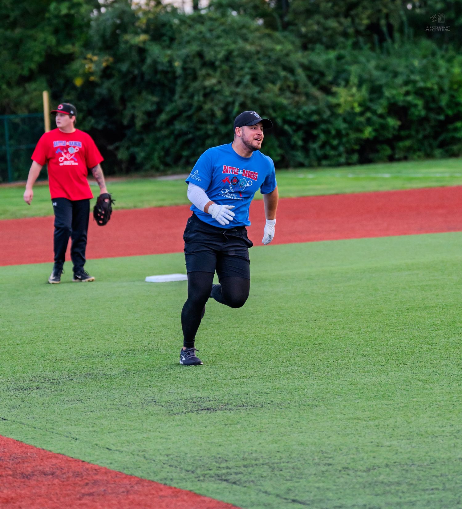 Two baseball players on a green field, one in blue shirt running bases, another in red standing near a base.
