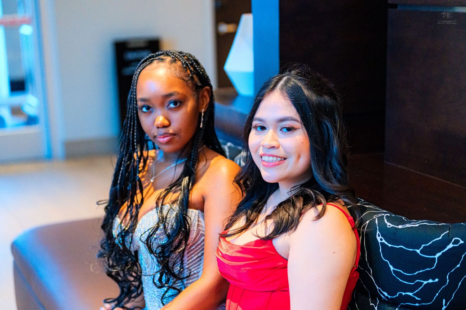 Two young women in formal dresses sitting together on a sofa posing for a photo.
