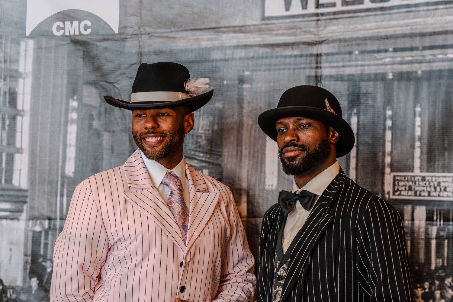 Two stylish men in pinstripe suits and fedora hats pose together at a vintage-themed event backdrop.