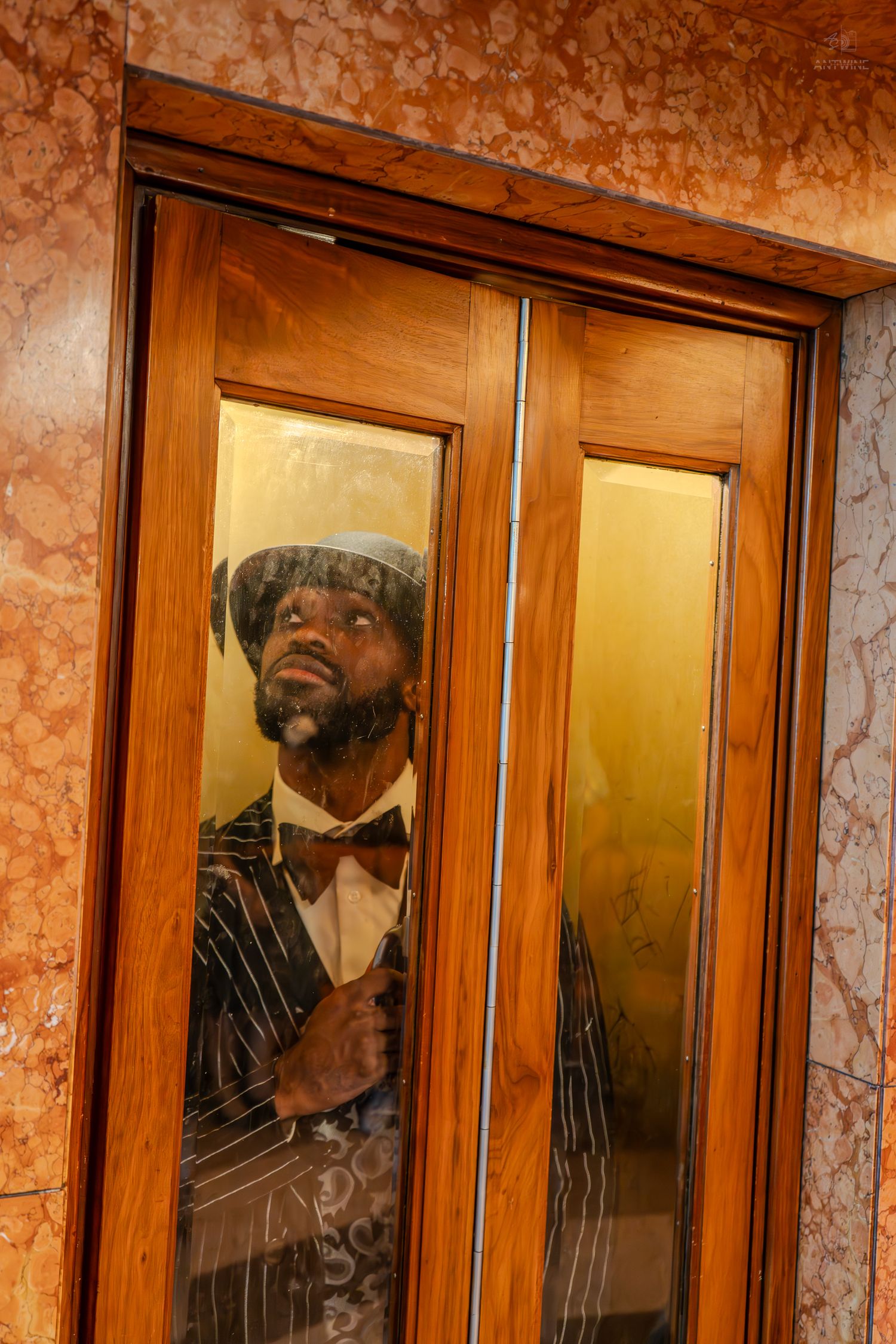 Stylish man in pinstripe suit and bow tie peers through glass elevator doors in ornate marble lobby.