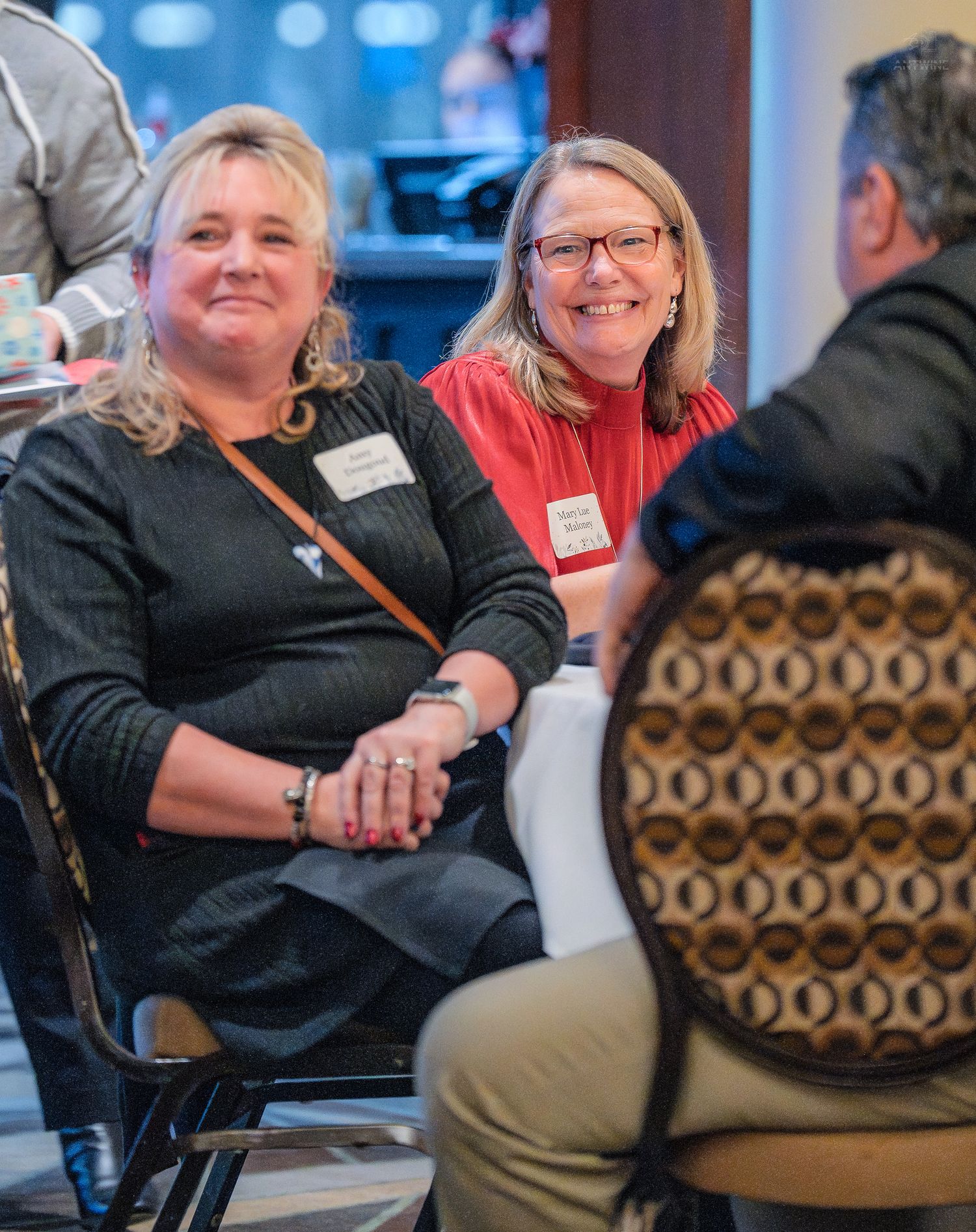 Two women wearing name tags smile warmly while seated at a networking event.