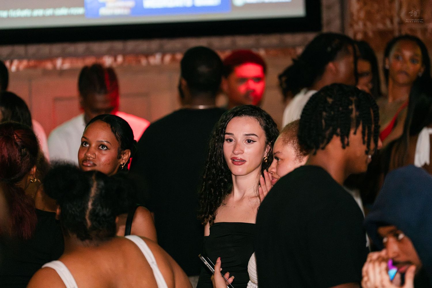 Young woman in a black dress gazes at the camera amid a lively crowded nightclub party.