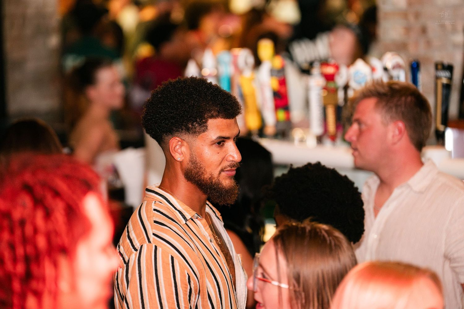 Young man in striped shirt standing near bar taps at a crowded nightclub.