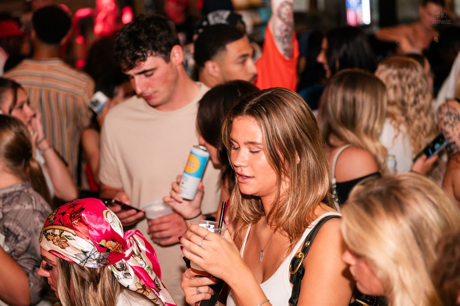 Young woman in white dress checking her phone while holding a drink at a busy bar.