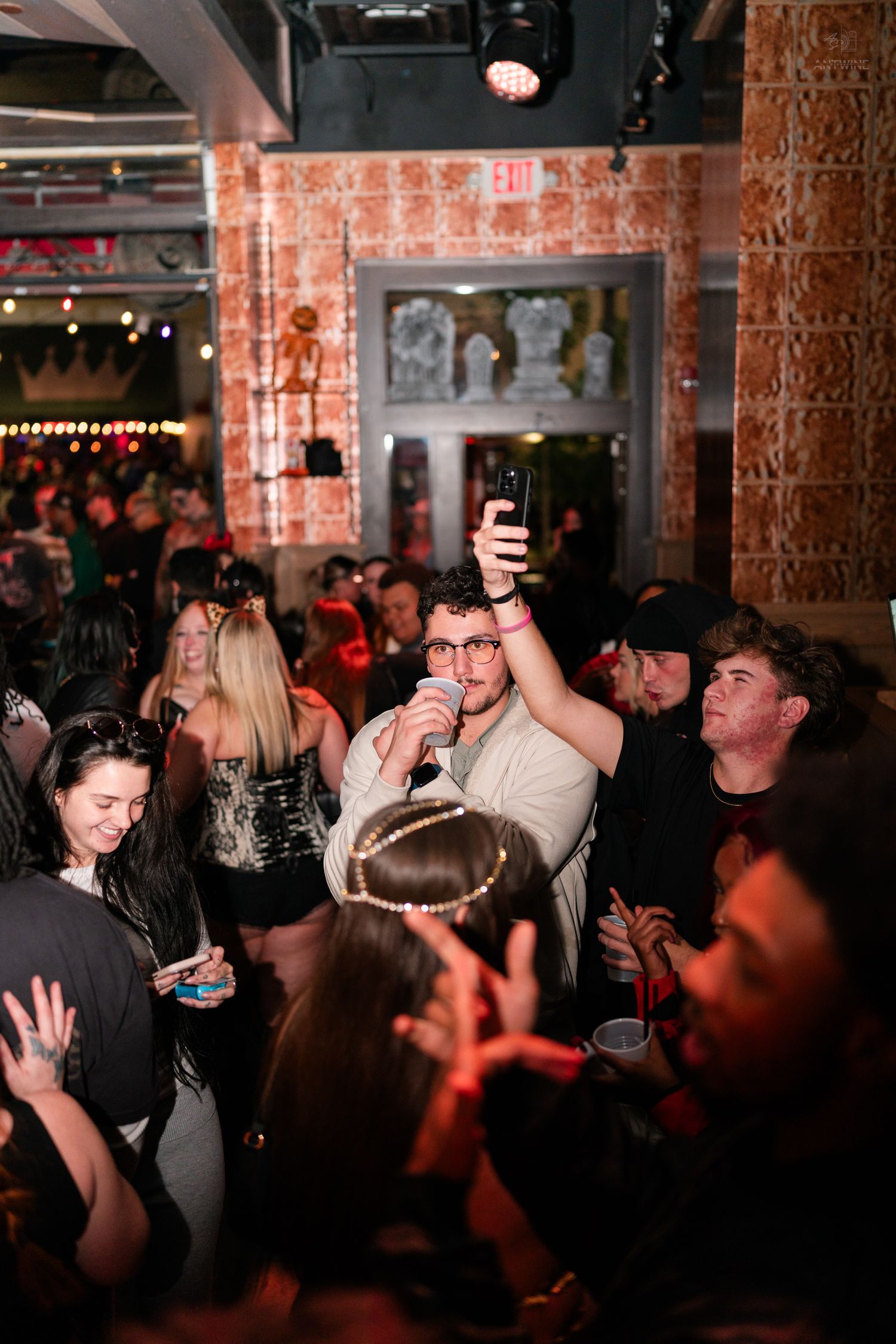 Man holds microphone and phone while performing for a large Halloween costume party crowd.