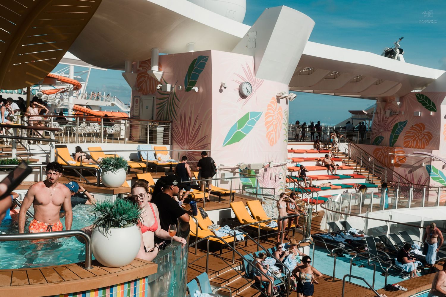 Wide view of a vibrant cruise ship pool deck with guests lounging under a clear blue sky.