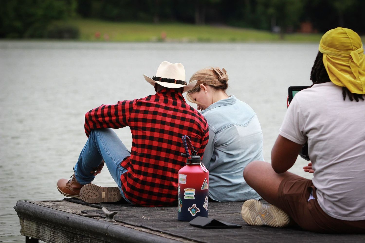 Three people sit together on a wooden dock by a lake at dusk, with a red water bottle beside them.