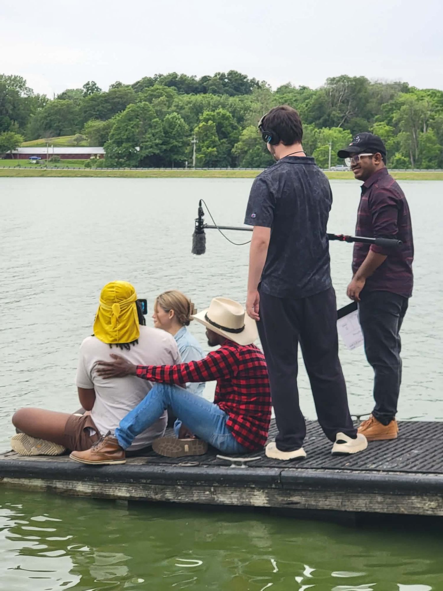 Film crew records actors sitting on a dock by a calm lake with green trees in the background.
