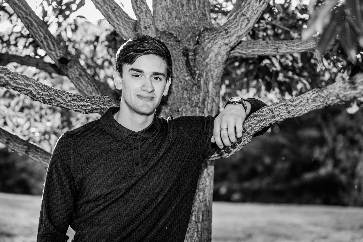 Young man leaning against a tree trunk outdoors, black and white portrait photo.