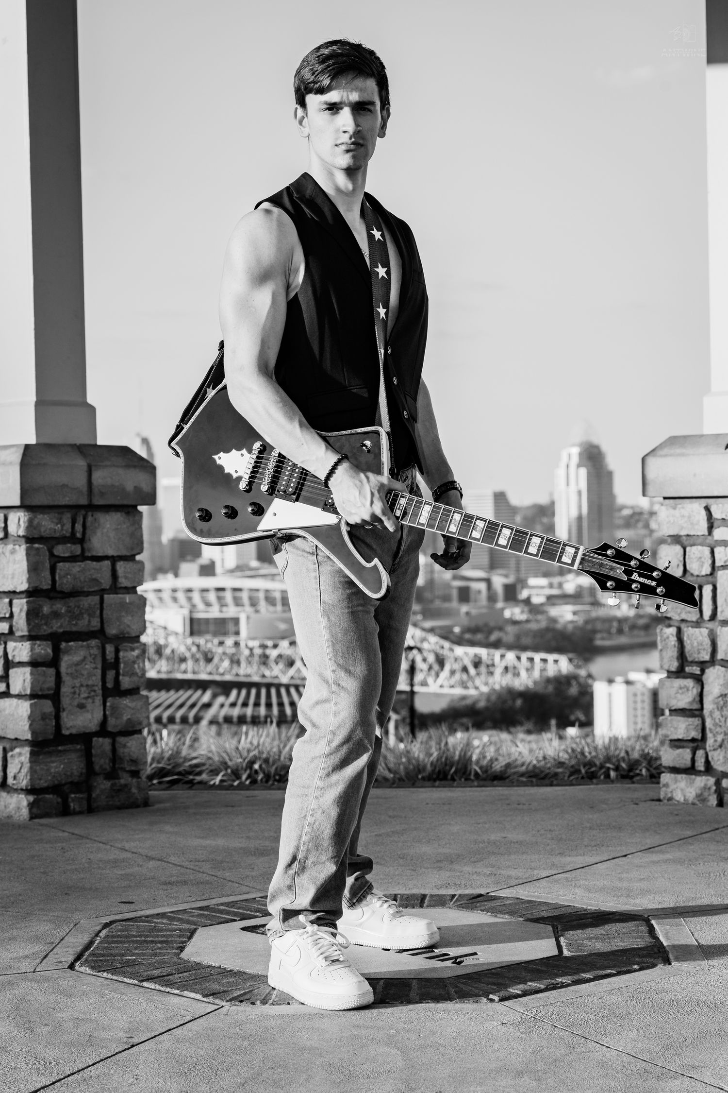 Young male guitarist holding electric guitar outdoors with city skyline background in black and white.