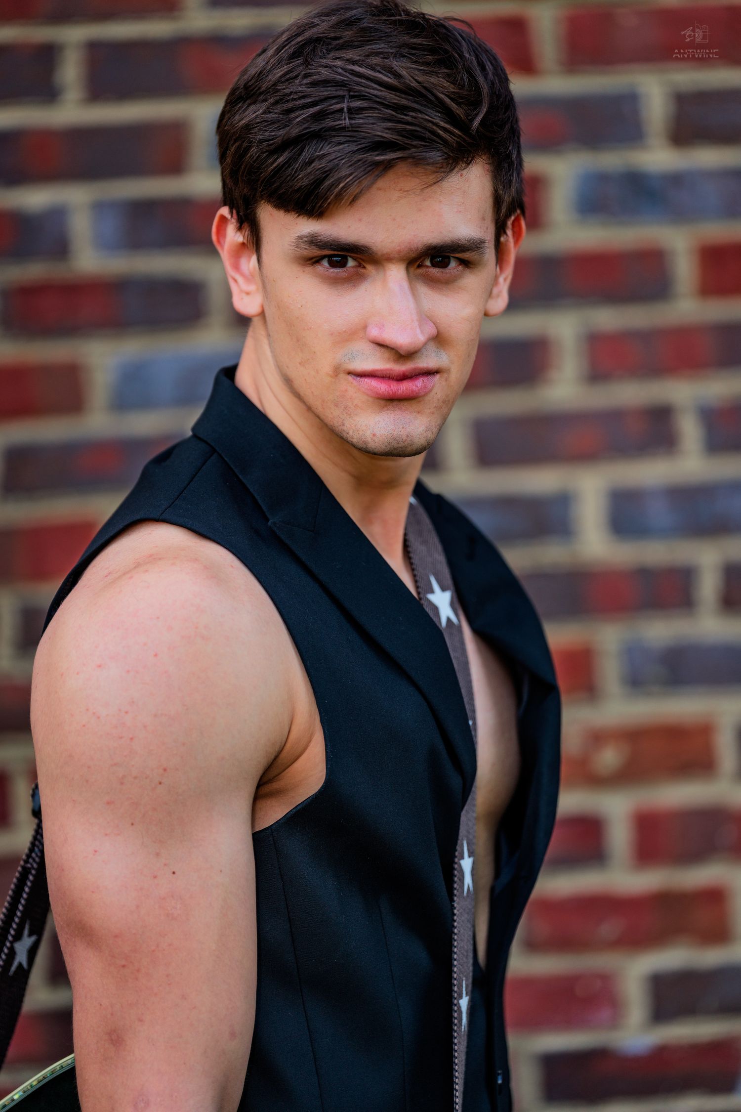 Young man in black sleeveless vest poses confidently against a brick wall outdoors.