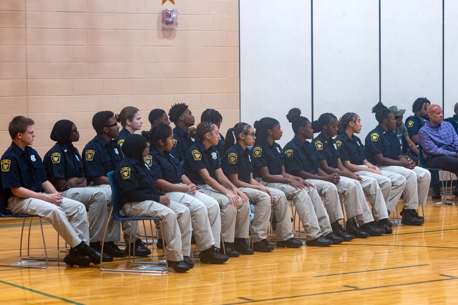 Youth cadets in dark uniforms and khaki pants seated in a row during a gymnasium ceremony.