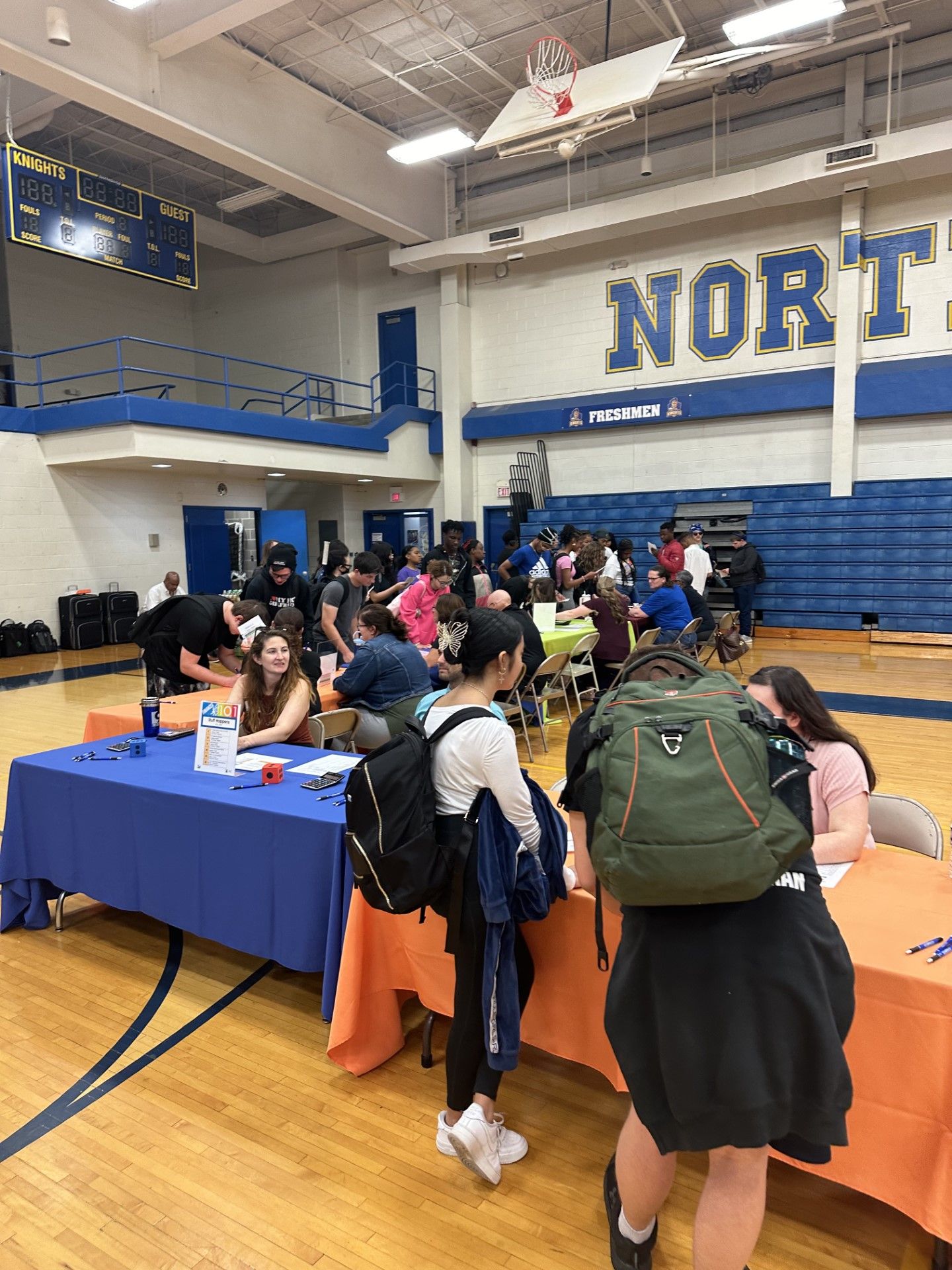 Students with backpacks gather at tables in a school gymnasium decorated in blue and white with NORTH on the wall.