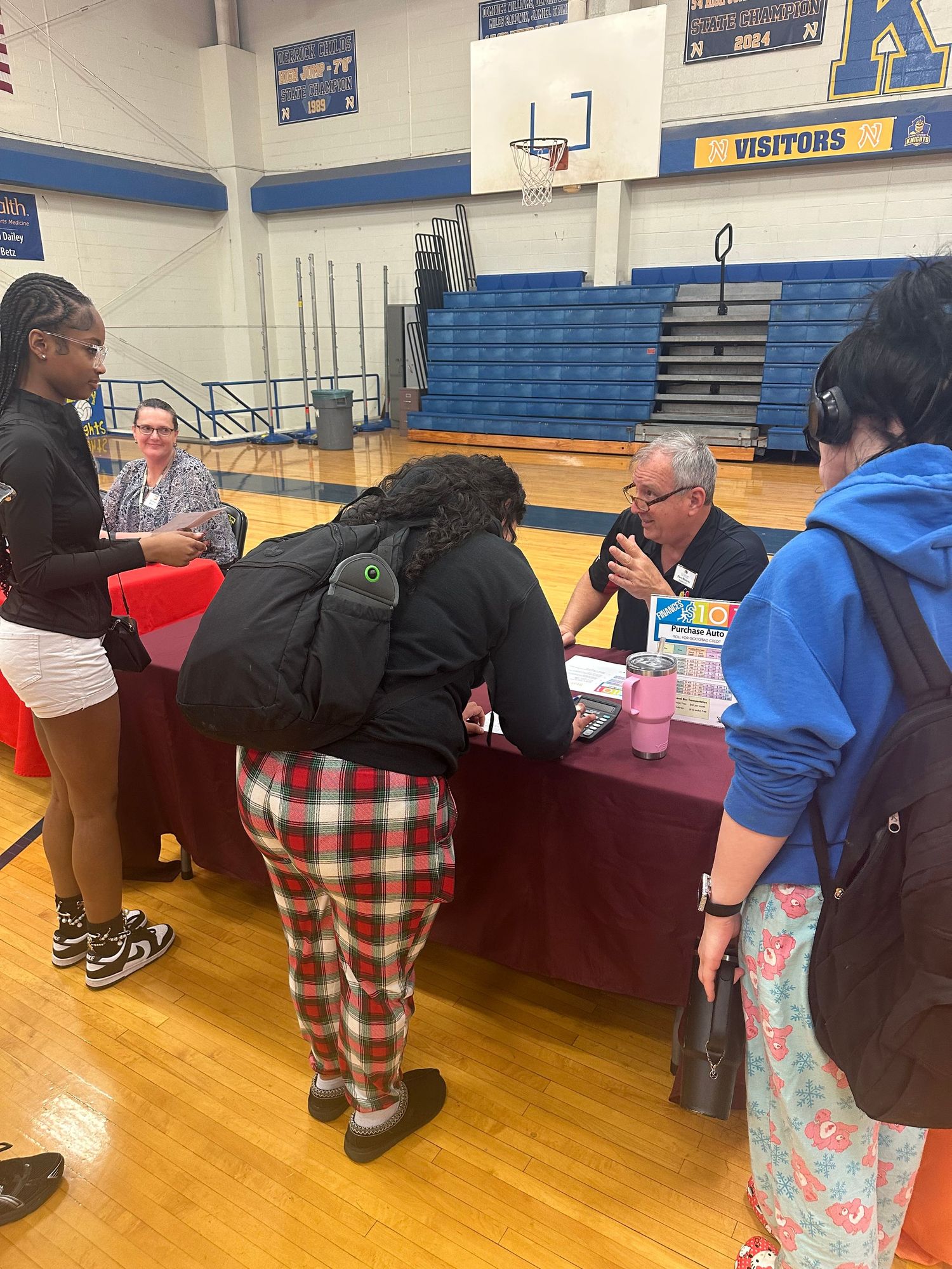 Students interact with a representative at a college fair table inside a school gymnasium.