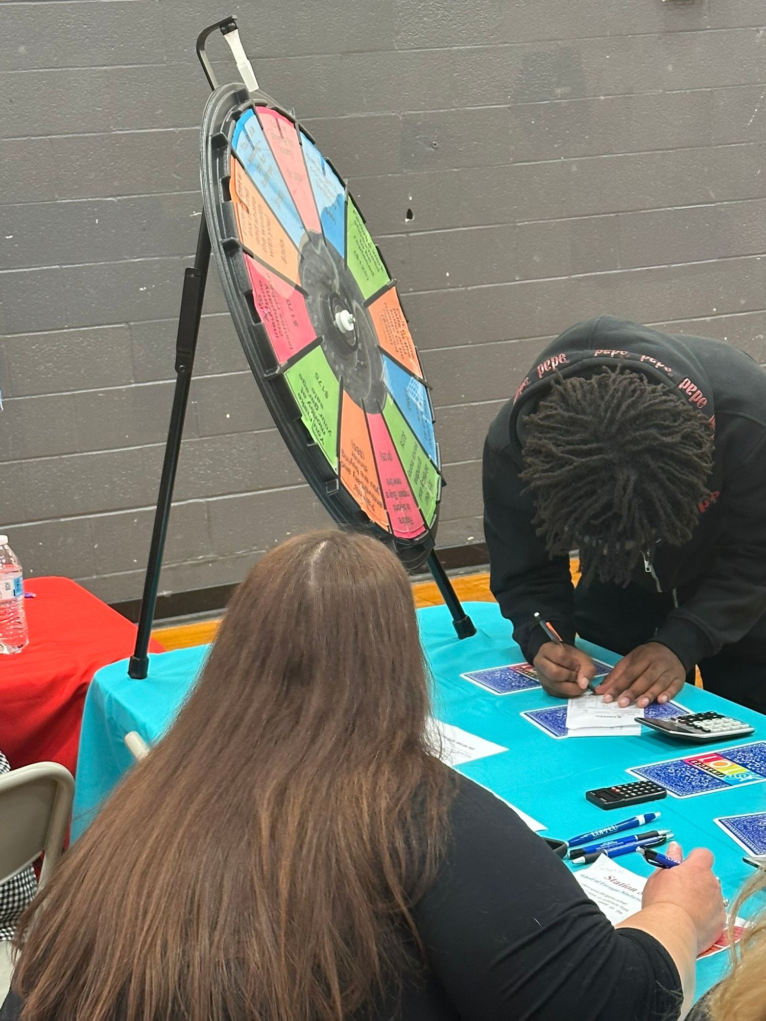 Student fills out form at colorful table with prize spin wheel at school fair event.