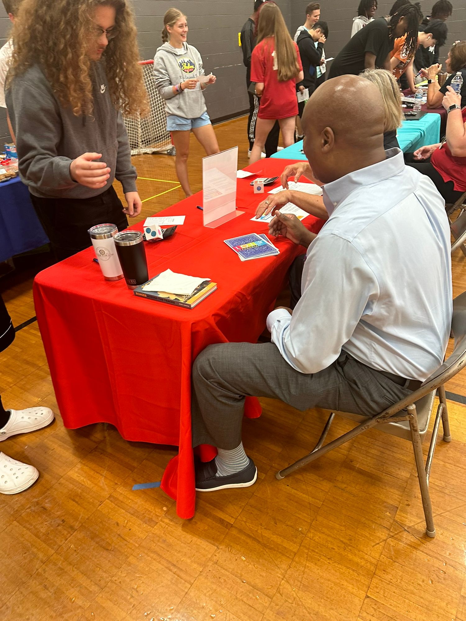 A man in a white shirt sits at a red table during a school career fair event in a gymnasium.