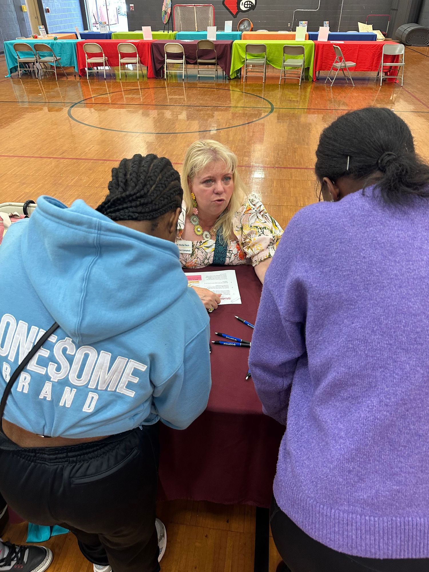 Three people having a meeting at a table in a school gymnasium, reviewing documents together.