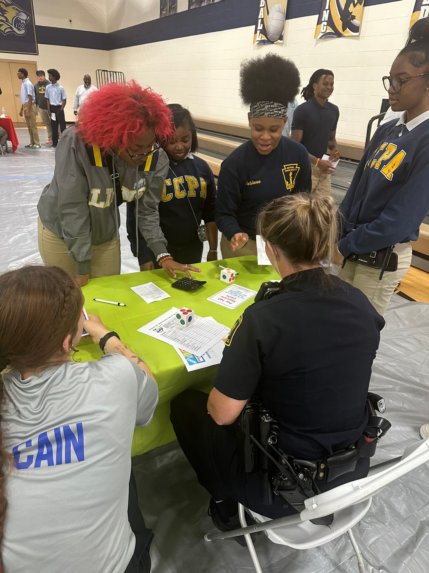 Students and officers interact at a green table during a school community engagement event in a gymnasium.
